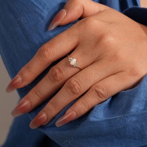 Puede incluir: Un anillo de oro con una piedra central de &oacute;palo ovalada, flanqueada por piedras preciosas m&aacute;s peque&ntilde;as y transparentes. El anillo est&aacute; engastado en una delicada banda de oro. La mano que lleva el anillo descansa sobre tela azul.