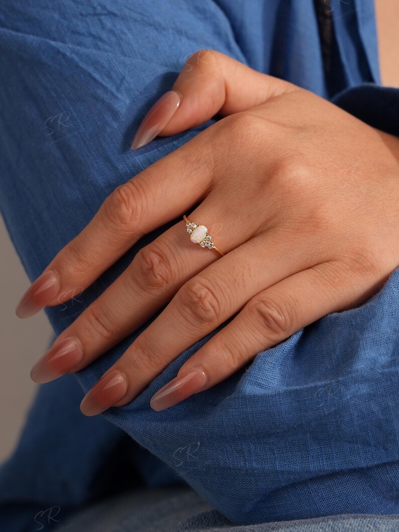 Puede incluir: Un anillo de oro con una piedra central de &oacute;palo ovalada, flanqueada por piedras preciosas m&aacute;s peque&ntilde;as y transparentes. El anillo est&aacute; engastado en una delicada banda de oro. La mano que lleva el anillo descansa sobre tela azul.