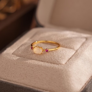 May include: A gold ring with an oval, off-white center stone and two small, round red stones. The ring is set in a gold band and is displayed in a gray ring box with a gold clasp. The ring is delicate and elegant.