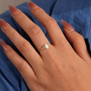 Puede incluir: Un anillo de oro con una piedra central de &oacute;palo ovalada, flanqueada por piedras de acento m&aacute;s peque&ntilde;as y transparentes. El anillo est&aacute; engarzado en una delicada banda de oro. La mano que lleva el anillo est&aacute; sobre un fondo azul.