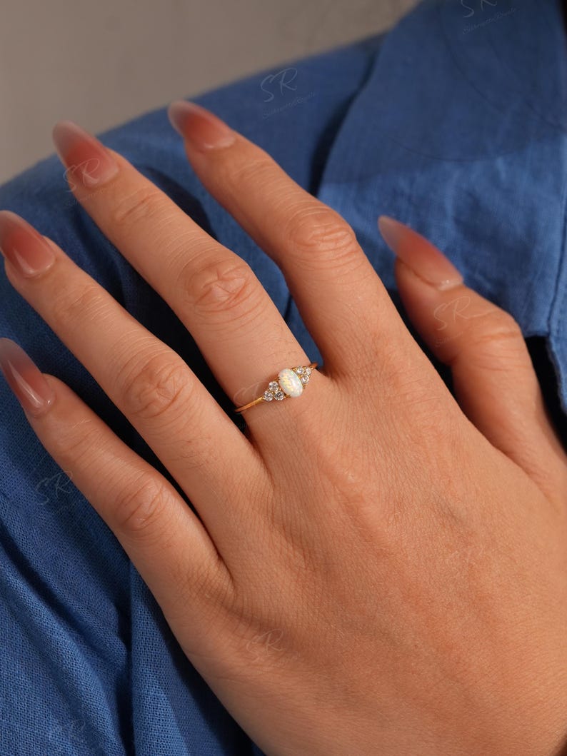 Puede incluir: Un anillo de oro con una piedra central de &oacute;palo ovalada, flanqueada por piedras de acento m&aacute;s peque&ntilde;as y transparentes. El anillo est&aacute; engarzado en una delicada banda de oro. La mano que lleva el anillo est&aacute; sobre un fondo azul.