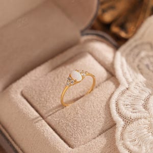 Puede incluir: Un anillo de oro con una piedra preciosa ovalada blanca en el centro, flanqueada por piedras claras m&aacute;s peque&ntilde;as. El anillo est&aacute; engastado en una banda de oro y se muestra en una caja beige. Es una joya.