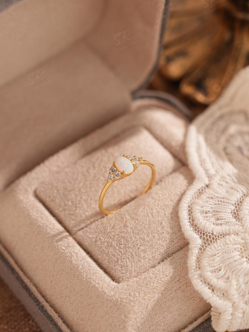 Puede incluir: Un anillo de oro con una piedra preciosa ovalada blanca en el centro, flanqueada por piedras claras m&aacute;s peque&ntilde;as. El anillo est&aacute; engastado en una banda de oro y se muestra en una caja beige. Es una joya.
