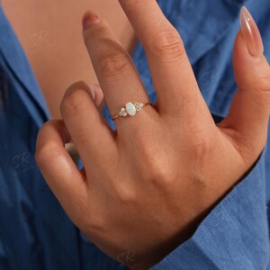 Puede incluir: Un anillo de oro con una piedra preciosa de &oacute;palo ovalada, flanqueada por piedras preciosas claras m&aacute;s peque&ntilde;as. El anillo tiene una banda delicada. El &oacute;palo es el punto focal, con las piedras m&aacute;s peque&ntilde;as que a&ntilde;aden brillo.