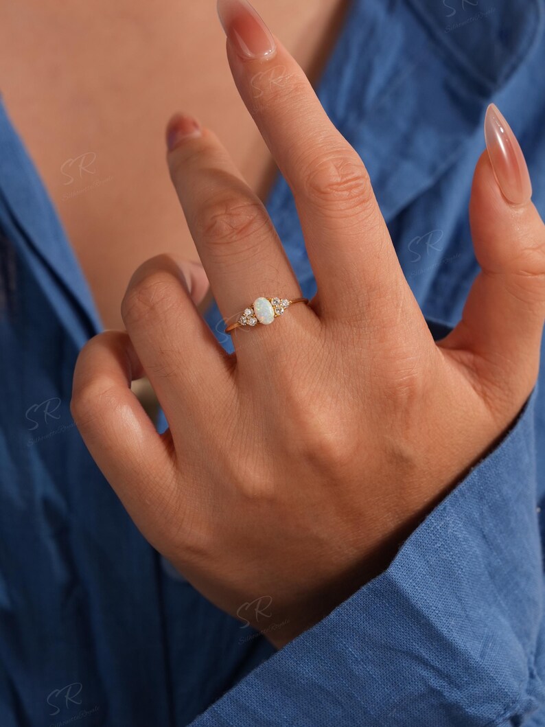 Puede incluir: Un anillo de oro con una piedra preciosa de &oacute;palo ovalada, flanqueada por piedras preciosas claras m&aacute;s peque&ntilde;as. El anillo tiene una banda delicada. El &oacute;palo es el punto focal, con las piedras m&aacute;s peque&ntilde;as que a&ntilde;aden brillo.