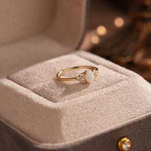 Puede incluir: Un anillo de oro con una piedra central de &oacute;palo ovalada, flanqueada por piedras preciosas claras m&aacute;s peque&ntilde;as. El anillo est&aacute; engastado en una banda de oro y se presenta en un joyero de terciopelo gris abierto.