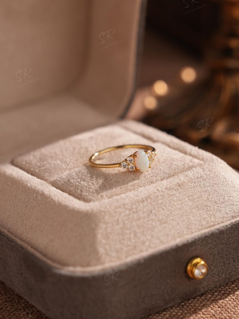Puede incluir: Un anillo de oro con una piedra central de &oacute;palo ovalada, flanqueada por piedras preciosas claras m&aacute;s peque&ntilde;as. El anillo est&aacute; engastado en una banda de oro y se presenta en un joyero de terciopelo gris abierto.