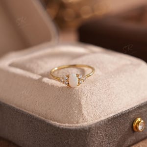 Puede incluir: Un anillo de oro con una piedra de &oacute;palo ovalada central, flanqueada por piedras transparentes m&aacute;s peque&ntilde;as. El anillo est&aacute; engastado en una banda de oro y presentado en una caja de joyer&iacute;a gris con interior beige. Una joya delicada.