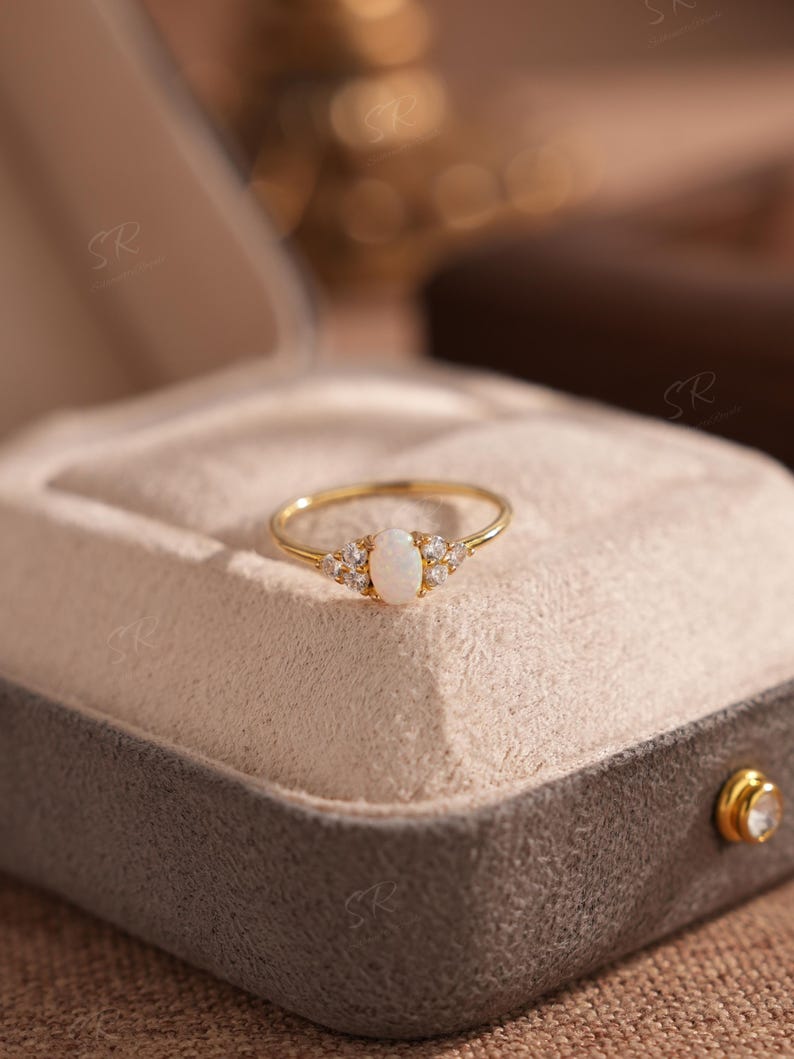 Puede incluir: Un anillo de oro con una piedra de &oacute;palo ovalada central, flanqueada por piedras transparentes m&aacute;s peque&ntilde;as. El anillo est&aacute; engastado en una banda de oro y presentado en una caja de joyer&iacute;a gris con interior beige. Una joya delicada.