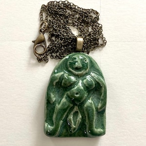 Puede incluir: Un collar con un colgante de cerámica verde que representa una figura femenina estilizada con una cara sonriente. El colgante está unido a una cadena de bronce.