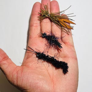 May include: Three fishing flies are displayed. One fly is made of brown and gold materials, resembling a cluster of needles. Two black flies with intricate details are also shown. The flies are designed for angling.