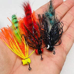 May include: Three colorful fishing flies with vibrant feathers and detailed hooks. The flies feature a variety of colors, including yellow, orange, red, green, and black. The hooks are black and the eyes are red and white.