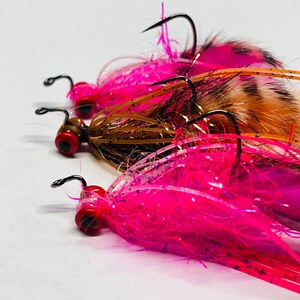 May include: Close-up of three fishing lures. Each lure features a black hook, a red eye, and a vibrant pink, brown, and orange feather design. The lures are arranged in a row, with the focus on the details of the craftsmanship.