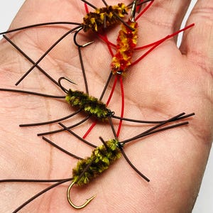 May include: Close-up of several artificial fishing flies. The flies have a fuzzy body in shades of yellow, brown, and red, with dark brown legs and a gold hook. Red and brown thread-like materials extend from the fly bodies.