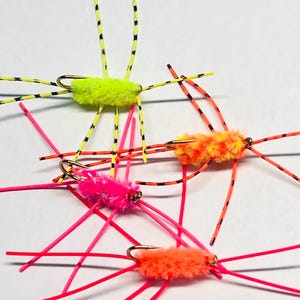 May include: Close-up of four colorful fishing flies. The flies have fuzzy bodies in neon yellow, orange, and pink, with gold hooks. Legs are made of thin, brightly colored wire, some with black stripes. These flies are designed for fly fishing.