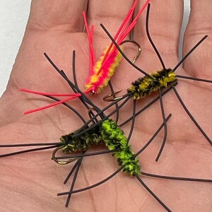 May include: Close-up of three colorful fishing flies. One fly is yellow and pink, another is yellow and black, and the third is green and black. Each fly has a gold hook and black wire legs, designed for fly fishing.
