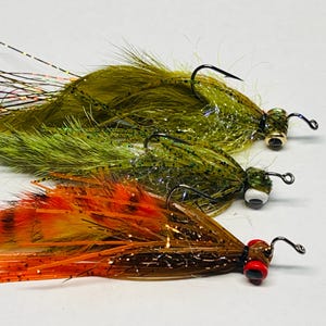 May include: Three fishing flies with black hooks. The top two flies are olive green with iridescent accents. The bottom fly is orange and brown with a red head. These flies are designed for catching fish.