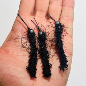 May include: Three black fishing flies with fuzzy bodies and thin, wire-like antennae. Each fly has a small, silver bead at the head. The flies are arranged on a light-colored surface.