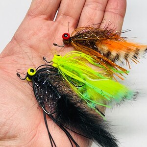 May include: Three colorful fishing flies with detailed construction. The flies feature vibrant colors including black, orange, brown, and green. Each fly has a hook and is designed for angling. The flies are designed to attract fish.