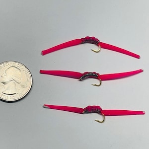 May include: Three bright pink fishing lures with gold hooks and dark thread detailing. The lures are arranged on a white surface next to a U.S. quarter coin for size comparison. The lures are designed to mimic worms.