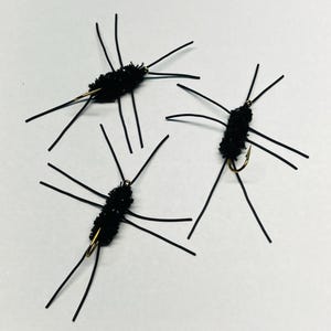 May include: Three black fishing flies with long, thin, black legs and a fuzzy body. The flies have a gold-colored hook and are designed to mimic insects. These flies are used for fly fishing.