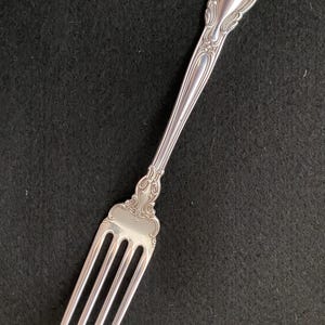 May include: Silver fork with ornate detailing. The fork features a decorative handle with a floral design and a four-tine head. The silverware is made of a shiny, reflective metal, likely silver or a silver-colored alloy. The fork is set against a dark background.