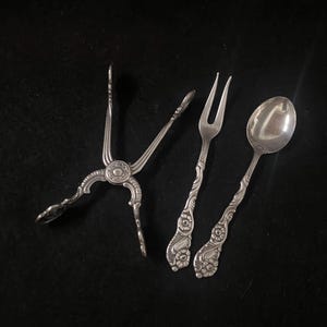 May include: An overhead shot of antique silver serving utensils on a black surface. The set includes a pair of tongs, a fork, and a spoon. Each piece has a decorative floral pattern on the handles.