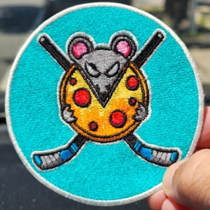 Pode incluir: Um remendo redondo azul bordado com um rato cinza segurando uma pizza amarela com pepperoni vermelho. O rato tem uma expressão ameaçadora e segura dois tacos de hóquei cruzados.