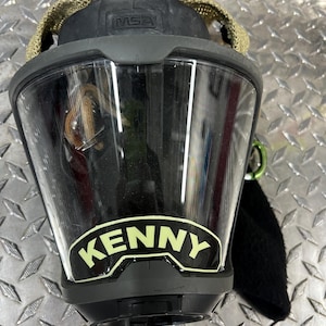 May include: A black and green firefighter's mask with a clear visor and the word "KENNY" printed in white on the front. The mask has a green strap and the letters "MSA" are visible on the top.