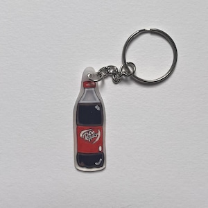 May include: A clear keychain with a silver metal ring and chain. The keychain features a red and black bottle with the word "Pepsi" printed on it.
