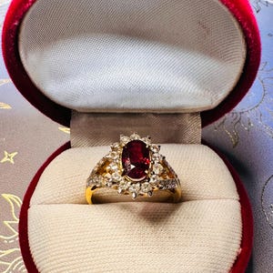 May include: A gold ring featuring a central oval-cut deep red gemstone surrounded by a halo of small round white stones. The band is crafted from yellow gold and the ring is displayed inside a red velvet-lined jewellery box with a cream interior.