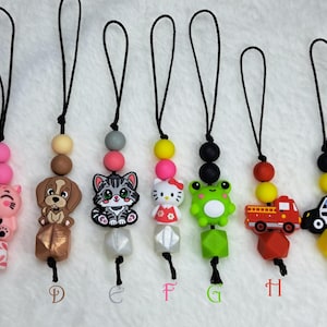 Puede incluir: Una colección de cordones de cuentas de silicona de colores con varios dijes. Los dijes incluyen pingüinos de dibujos animados, un cerdo rosa, un perro marrón, un gato gris, Hello Kitty, una rana verde, un camión de bomberos rojo, un coche de policía, una abeja y un pato amarillo.