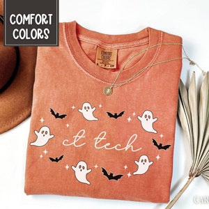May include: A burnt orange t-shirt with a round neck featuring a design of white ghosts, black bats, and the text "ct tech" in white. The shirt has a tag that says "Comfort Colors". A gold necklace and a brown hat are also in the image.