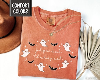 May include: A coral-colored t-shirt with the words "physical therapist" in white script, surrounded by white ghost and black bat graphics. The shirt has a crew neck and short sleeves. The words "Comfort Colors" are printed on the shirt's tag.