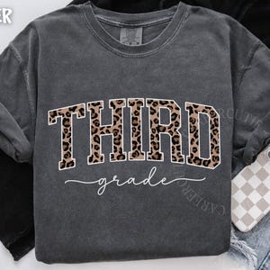 May include: Dark gray sweatshirt with the word "THIRD" in leopard print and "grade" in cursive. The shirt is a comfortable, casual style, perfect for back-to-school or everyday wear.