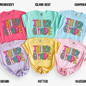 May include: A collection of six colorful t-shirts, each in a different pastel shade: pink, light blue, yellow, lavender, light green, and pink. Each shirt features the words "THIRD GRADE" in a playful, multi-colored design with stars and a lightning bolt.