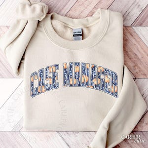 May include: A beige sweatshirt with the text "CASE MANAGER" in a blue and orange floral design. The sweatshirt is folded on a wooden surface. The brand label "GILDAN" is visible.