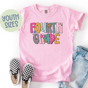 K&ouml;nnte beinhalten: Ein hellrosa Kinder-T-Shirt mit den Worten "FOURTH GRADE" in bunten Buchstaben im Retro-Stil. Das Shirt wird mit einer blauen Jeans und schwarz-wei&szlig; karierten Slip-On-Schuhen kombiniert. Eine hellgr&uuml;ne Blase mit der Aufschrift "YOUTH SIZES".