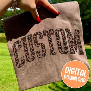 May include: A brown t-shirt with the word "CUSTOM" in large, bold letters with a leopard print design. The shirt is held up by a hand with red nail polish. An orange circle with the words "DIGITAL DOWNLOAD" is in the bottom right corner.