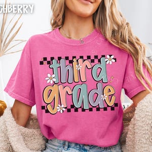 Op de afbeelding: Een felroze t-shirt met de tekst "third grade" in een kleurrijk, retro lettertype. De tekst staat tegen een zwart-witte geblokte achtergrond, met madeliefjesaccenten. Het shirt heeft een casual stijl.