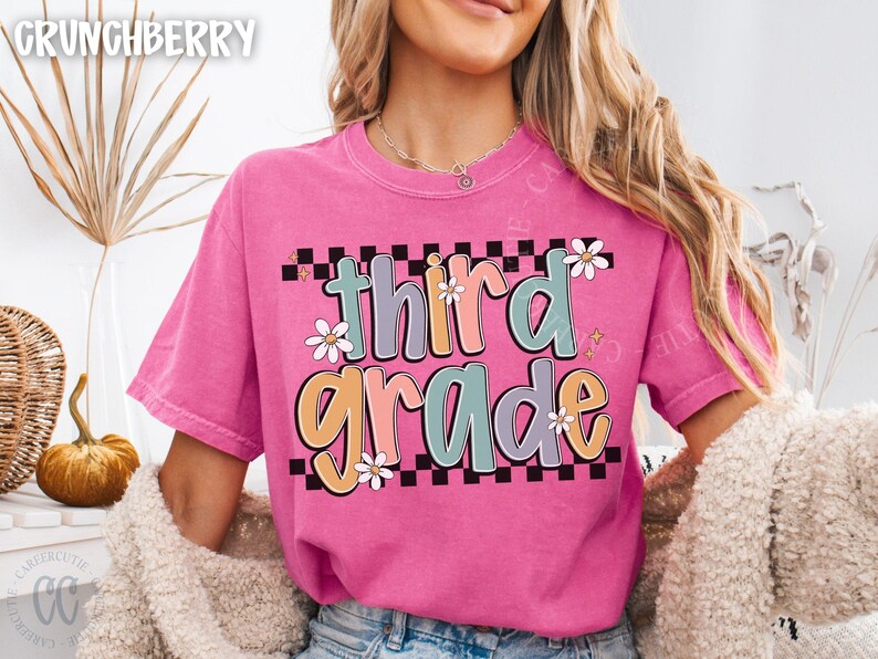 Op de afbeelding: Een felroze t-shirt met de tekst "third grade" in een kleurrijk, retro lettertype. De tekst staat tegen een zwart-witte geblokte achtergrond, met madeliefjesaccenten. Het shirt heeft een casual stijl.