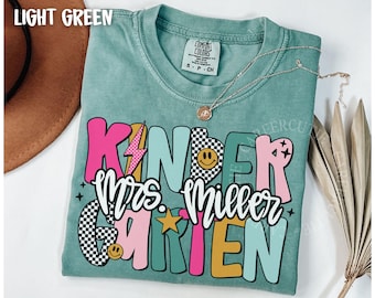 Puede incluir: Camiseta verde claro con las palabras "Kinder Mrs. Miller Garten" en una fuente colorida y juguetona. El diseño incluye un rayo, estrellas y una carita sonriente. Un collar plateado con un colgante está en la camiseta.