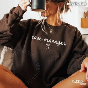 May include: Dark chocolate-colored sweatshirt with the words "case manager" in white script. A small bow graphic is below the text. The sweatshirt has long sleeves and a crew neck. The brand name "CAREER cutie" is printed on the bottom of the sweatshirt.