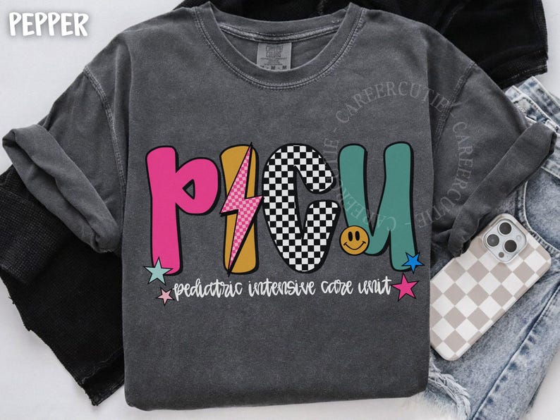 May include: Dark gray sweatshirt with the letters "PICU" in pink, yellow, black and white checkered, and teal. The text "pediatric intensive care unit" is below the letters. A phone with a checkered case is visible.