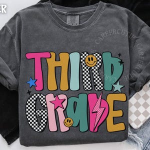 May include: Dark gray t-shirt with a colorful "THIRD GRADE" graphic. The design features the text in various colors, including pink, teal, and yellow, with stars, a lightning bolt, and smiley faces. The shirt is displayed on a denim background.