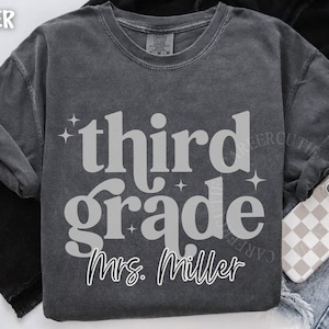 May include: Dark grey third grade t-shirt with the name "Mrs. Miller" in a script font. The shirt has a vintage, washed look. A phone with a checkered case and denim jeans are in the background.