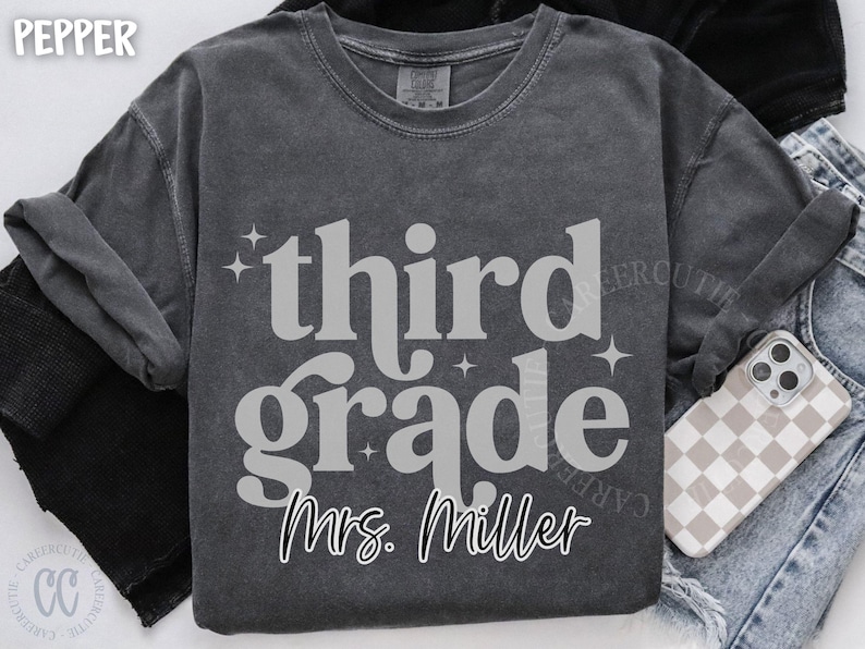 May include: Dark grey third grade t-shirt with the name "Mrs. Miller" in a script font. The shirt has a vintage, washed look. A phone with a checkered case and denim jeans are in the background.