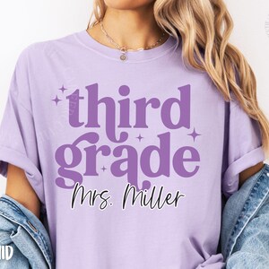 May include: A lilac t-shirt featuring the words "third grade" in a large, purple, retro font, with the name "Mrs. Miller" in cursive. The shirt has a relaxed fit and short sleeves.
