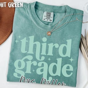 May include: Light green t-shirt with the words "third grade" in a light green font, with the name "Mrs. Miller" below. A gold necklace with a pendant is on the shirt. The shirt is a Comfort Colors brand.