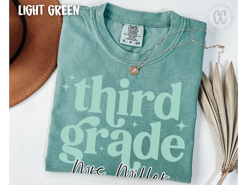 May include: Light green t-shirt with the words "third grade" in a light green font, with the name "Mrs. Miller" below. A gold necklace with a pendant is on the shirt. The shirt is a Comfort Colors brand.